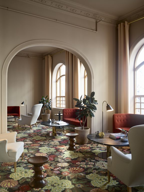 Fotografiet er tatt i hotellets lounge, gulvet er belagt med et blomstrete teppe. Veggene er malt i lys beige tone. Moderne møbler som tar opp fargene fra interiøret er plassert ut i rommet.
