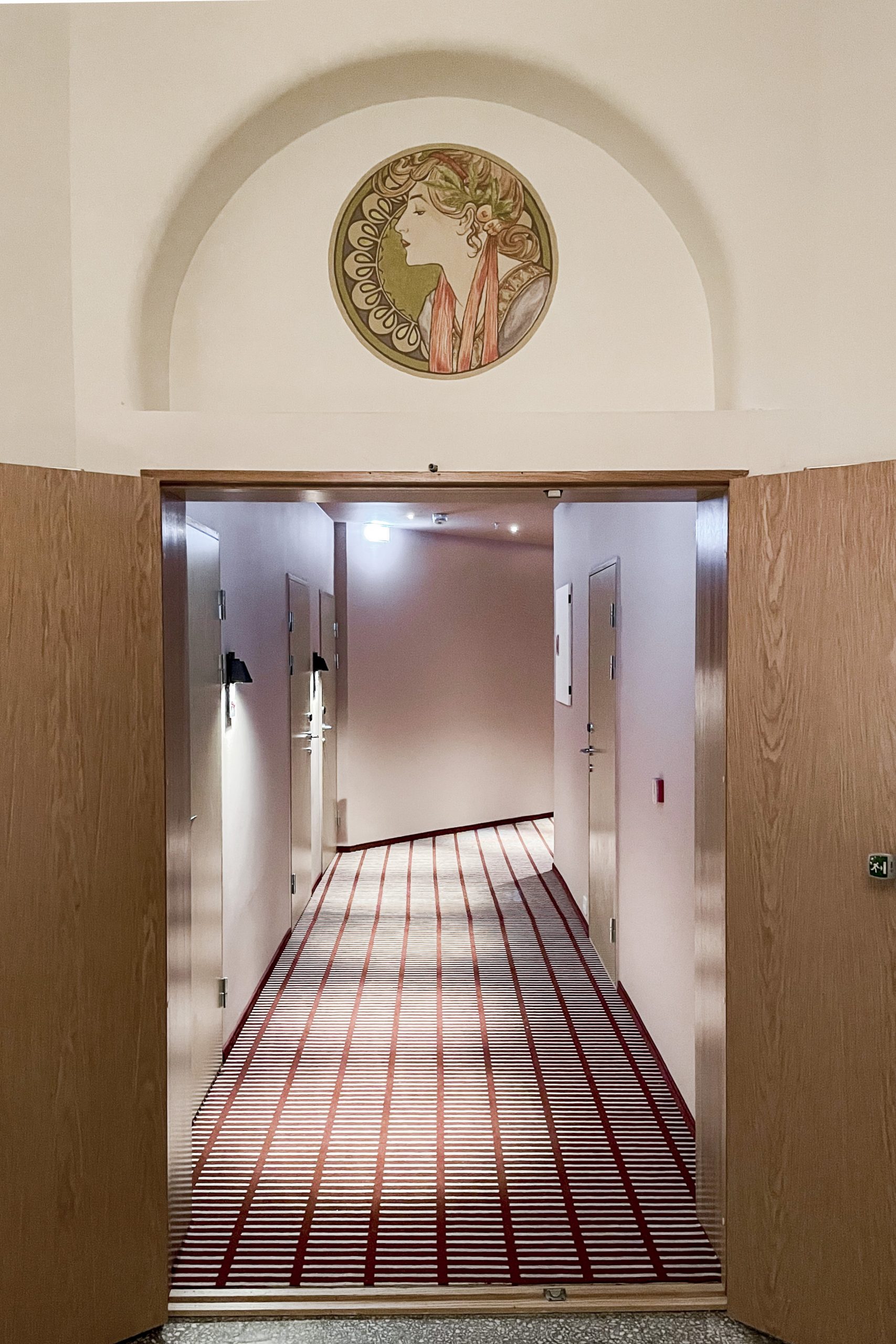 Fotografi som viser en av hotellkorridorene. Gulvet er belagt med et rutete teppe. I bildets forgrunn ser man et opprinnelig maleri i jugendstil plassert over døren.