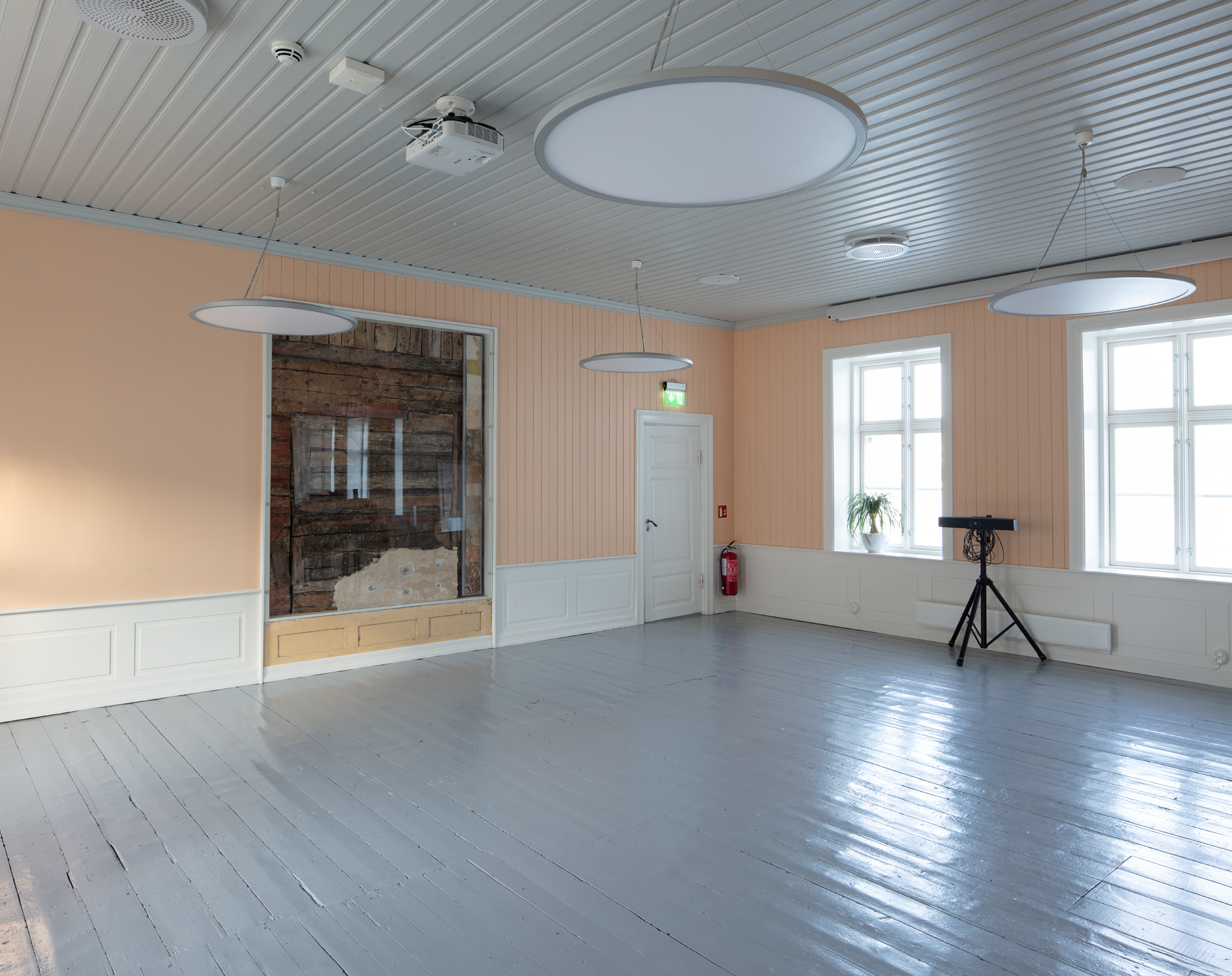 Bildet viser et større rom kledd i trepanel med malt tregulv. Veggene er malt i en lakserosa farge, og i midten av bildet ser man at det er felt inn et vindu som viser de originale veggene i brunt tømmer, bak den rosa trekledningen.