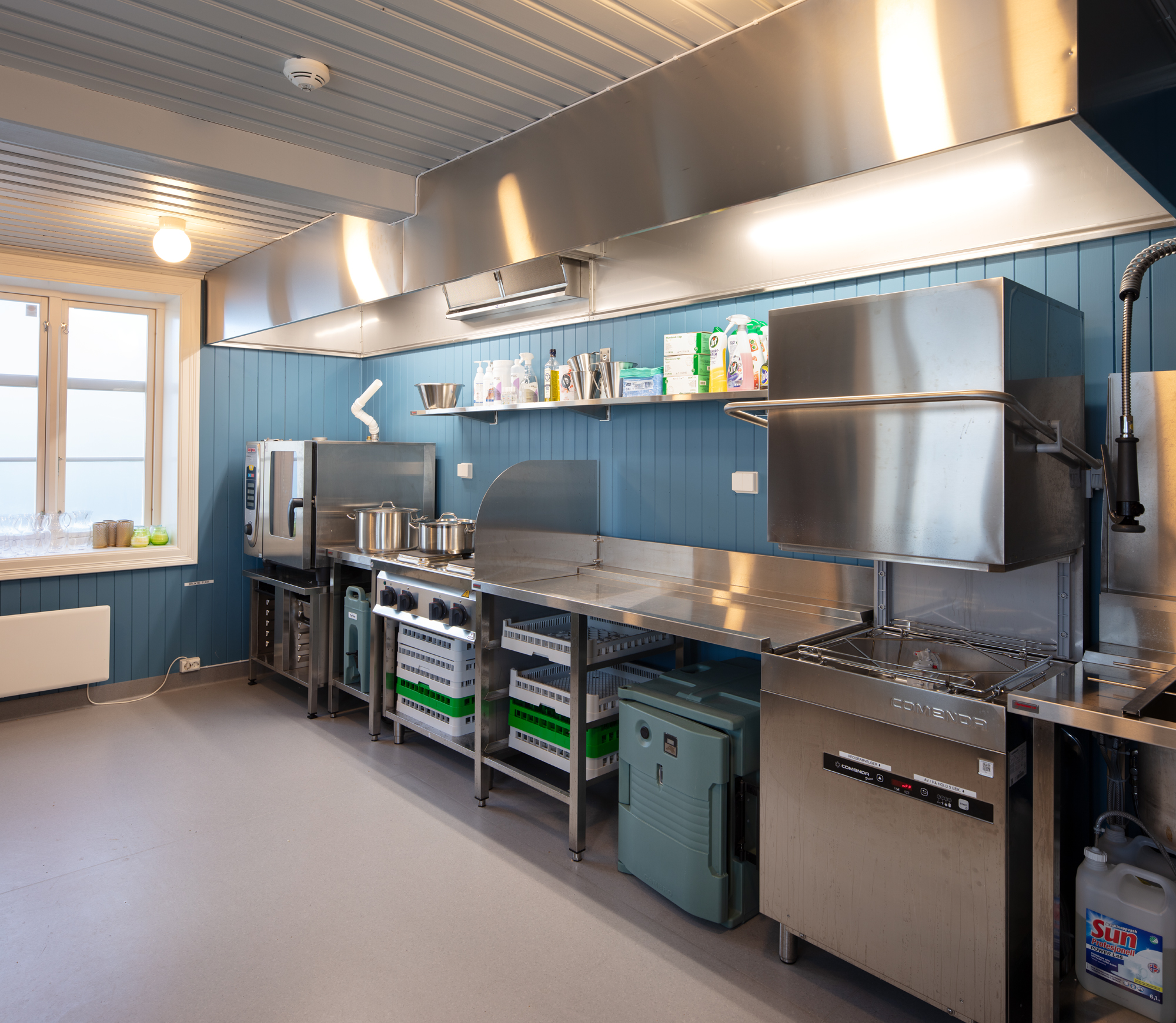 Bildet viser et kjøkken med stålfronter og overflater, med en gammeldags trevegg i mellomblå farge.